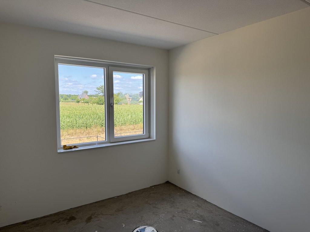 Witte slaapkamer in een nieuwe woning met gladde vliesmuren en een raam dat uitkijkt op een maïsveld