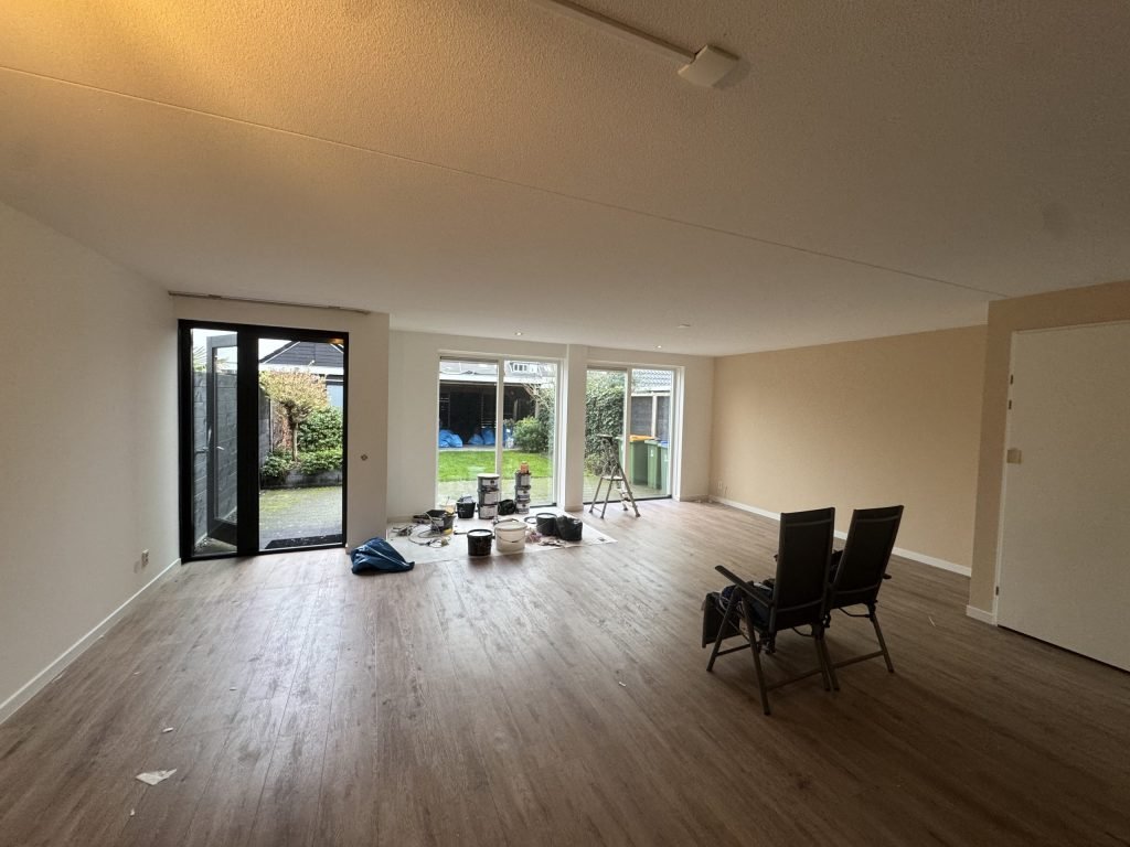 Ruime woonkamer in een bestaand huis met wit en zandkleurig gesausde muren, een trapje en verfemmers bij de tuindeuren
